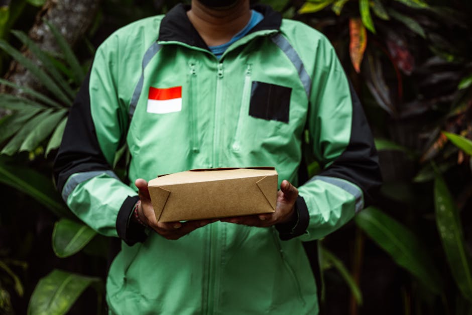 A person wearing a green and black jacket with an Indonesian flag patch and black accents is holding a rectangular, light-brown paper parcel wrapped with folded edges, in front of a lush background of