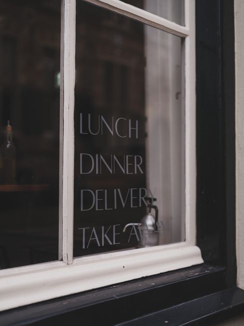 A close-up of a window with a white-painted wooden frame, reflecting a faint outdoor scene due to the glass. On the glass, words are listed vertically in white capital letters: 'LUNCH,' 'DINNER,' 'DEL