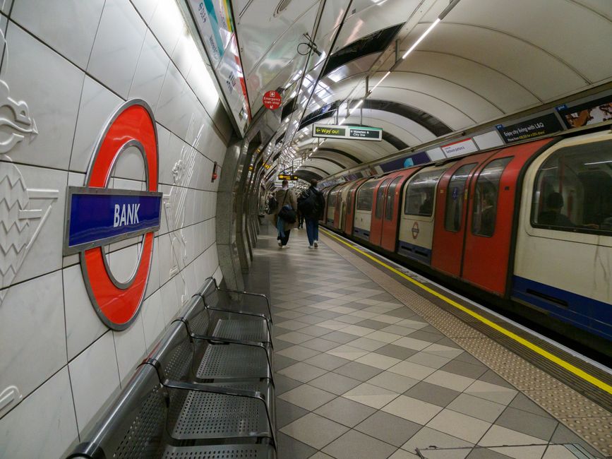 An underground platform at a London tube station with tiled walls and a large roundel sign featuring the word 'BANK' in blue. The platform has a row of black metal benches and a tiled floor arranged i