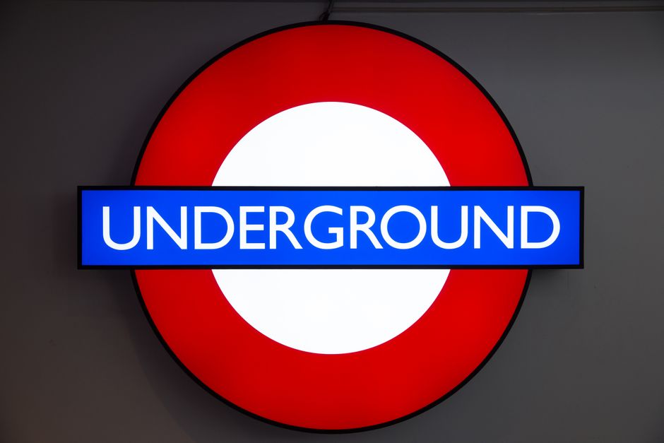Close-up of a London Underground sign featuring a red circle with a white central disc and a blue bar across the middle displaying the word 'UNDERGROUND' in white uppercase letters. The sign is mounte