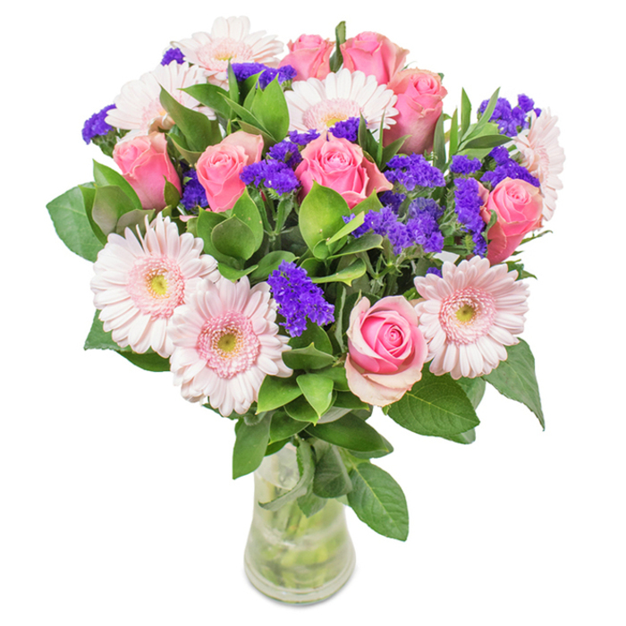 This radiant floral arrangement, designed with the Woodside Park community in mind, is artfully presented in a clean glass vase set against a pale, inviting backdrop. The centrepiece draws the eye with its harmonious medley: seven to nine pink roses, each unfurling in classic spirals with some still tightly budded, showcase a nuance of pinks from blush to true-rose hues. Nestled beside them, five to seven pale pink gerbera daisies with their signature large, round heads and creamy, almost white centres add lightness and contrast, recalling the cheerful blooms seen in front gardens along Torrington Park. Accented with vivid purple statice clustered throughout, the deeper tones introduce pops of texture and depth-much like glimpses of violets in a shaded Finchley woodland. Lush, deep green foliage spills around the blooms, lending vibrancy and grounding the arrangement with fullness and a just-picked freshness. The clear vase, with stems visible through sparkling water, enhances the sense of renewal and local abundance, perfect for a birthday gift, an anniversary gesture, or as a decorative centrepiece for a Woodside Park office or home. The bouquet's overall ambiance conveys joy, celebration, and a gentle nod to the natural landscapes surrounding this North London neighbourhood.